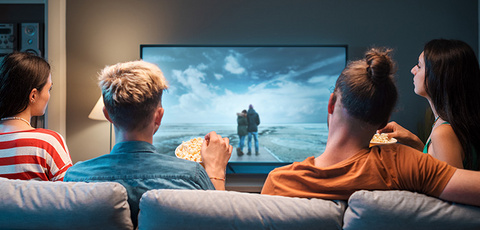 Quatre personnes sont assises sur un canapé, mangent du pop-corn et regardent ensemble un film sur une grande télévision.
