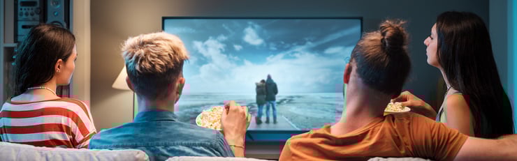 Quatre personnes sont assises sur un canapé, mangent du pop-corn et regardent ensemble un film sur une grande télévision.