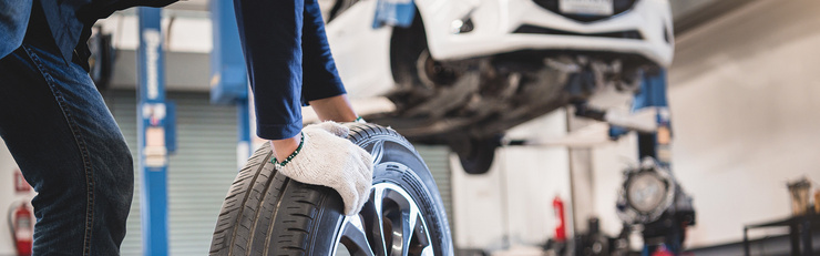 Une personne portant des gants de travail soulève un pneu de voiture dans un atelier, tandis qu'un véhicule est posé sur un pont élévateur.