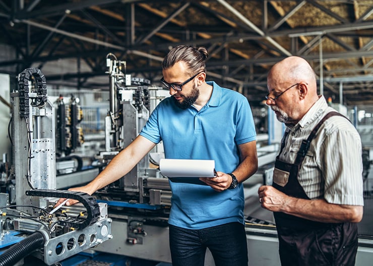 Deux personnes se trouvent dans un hangar industriel et inspectent ensemble une machine, tandis qu'une autre personne prend des notes sur un bloc-notes.