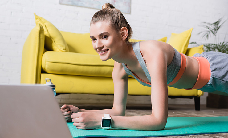 Une femme s'entraîne en position de planche sur un tapis de fitness à la maison et regarde un ordinateur portable.