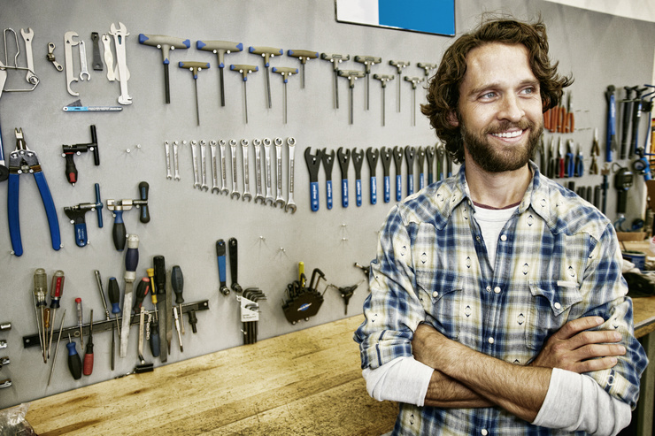 Un homme portant une chemise à carreaux se tient dans un atelier devant un mur avec des outils.