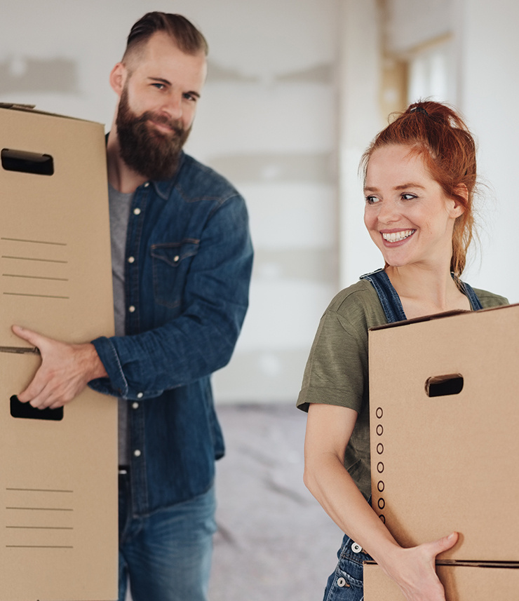 Un homme et une femme portent des cartons de déménagement en souriant dans une pièce en rénovation.