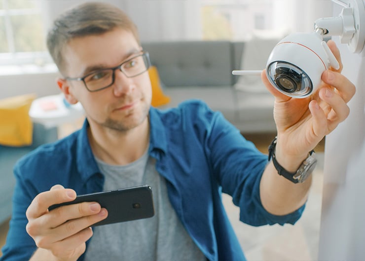 Un homme fixe une caméra de surveillance blanche sur un mur intérieur tout en tenant un smartphone dans la main.