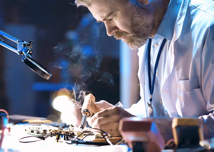 Un technicien travaille avec concentration sur un circuit imprimé électronique à l'aide d'un fer à souder, tandis qu'une fine fumée s'élève.