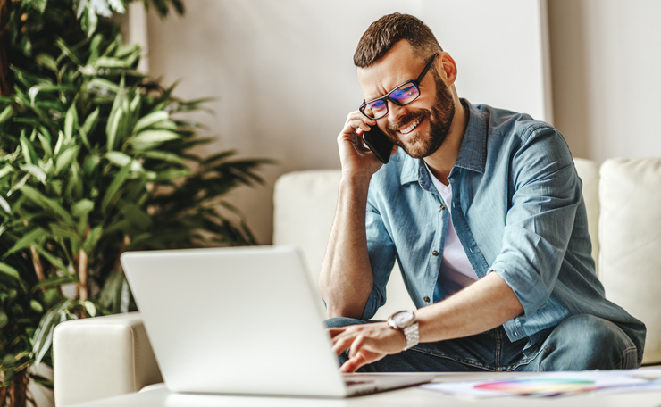 Un homme souriant avec des lunettes est assis sur un canapé, parle au téléphone et travaille sur un ordinateur portable.