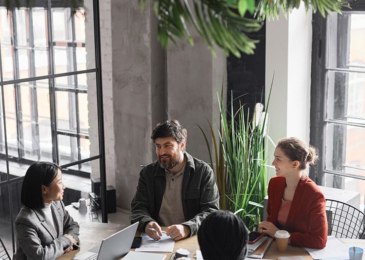 Vier Personen sitzen an einem Tisch in einem hellen Büro und führen ein gemeinsames Meeting mit Laptop und Unterlagen.