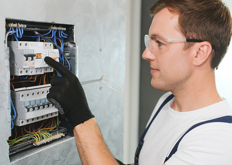 Un électricien portant des lunettes de protection et des gants inspecte un tableau électrique avec des câbles et des interrupteurs apparents.