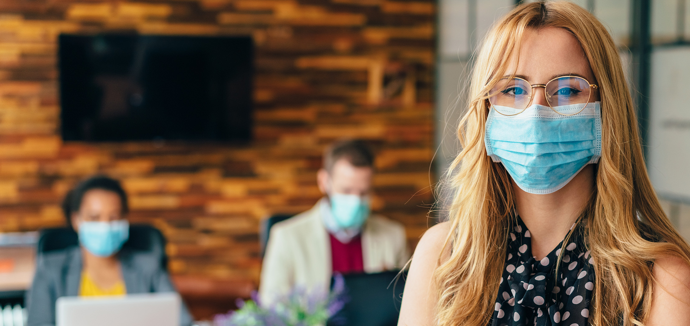 Une femme portant des lunettes et un masque médical se tient au premier plan d'un bureau, tandis que deux autres personnes masquées sont assises à l'arrière-plan.