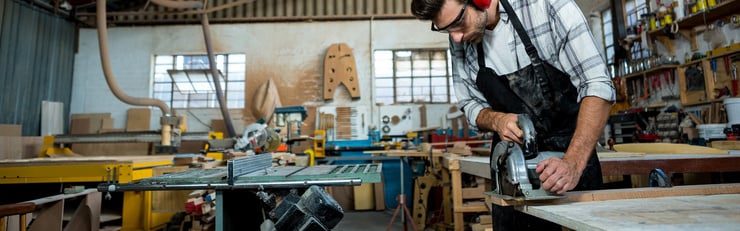 Une personne équipée d'une protection auditive et de lunettes de sécurité travaille dans un atelier sur une plaque de bois à l'aide d'une scie circulaire à main.