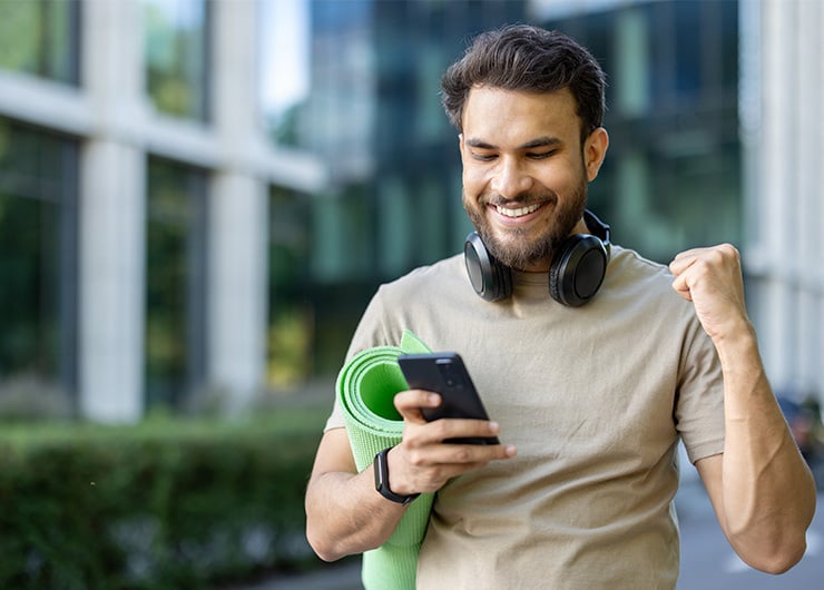 Un homme debout dehors avec des écouteurs, un téléphone portable et un tapis, levant joyeusement le poing en l'air.