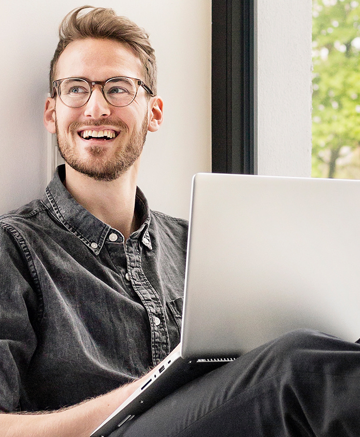Homme avec des lunettes assis près de la fenêtre travaillant en souriant sur un ordinateur portable.
