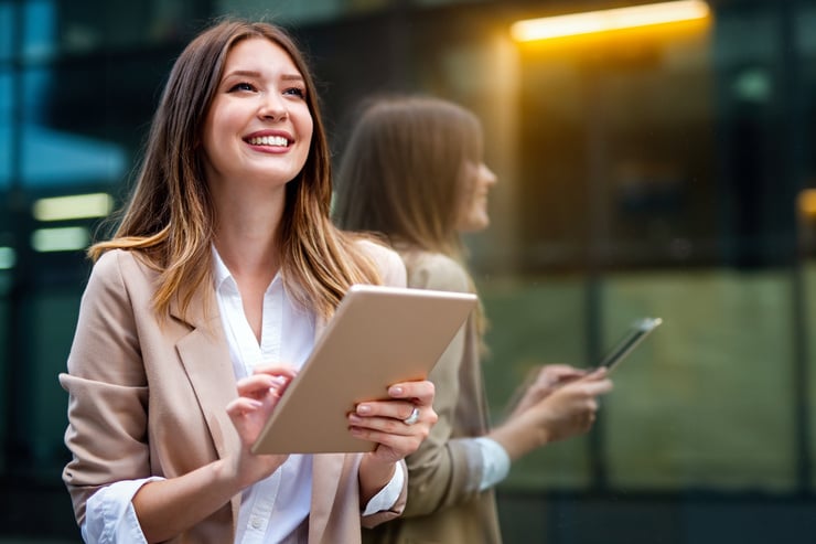 Deux jeunes femmes se tiennent devant un bâtiment moderne, l'une tient une tablette et sourit, l'autre regarde son smartphone.