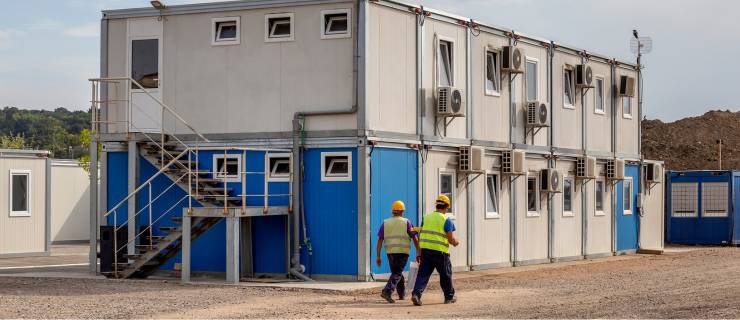 Deux ouvriers en gilets de sécurité et casques marchent devant un bâtiment modulaire à deux étages sur un chantier.