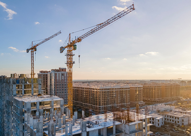 De grandes grues de chantier surplombent un complexe résidentiel en construction composé de plusieurs immeubles de grande hauteur, au coucher du soleil.