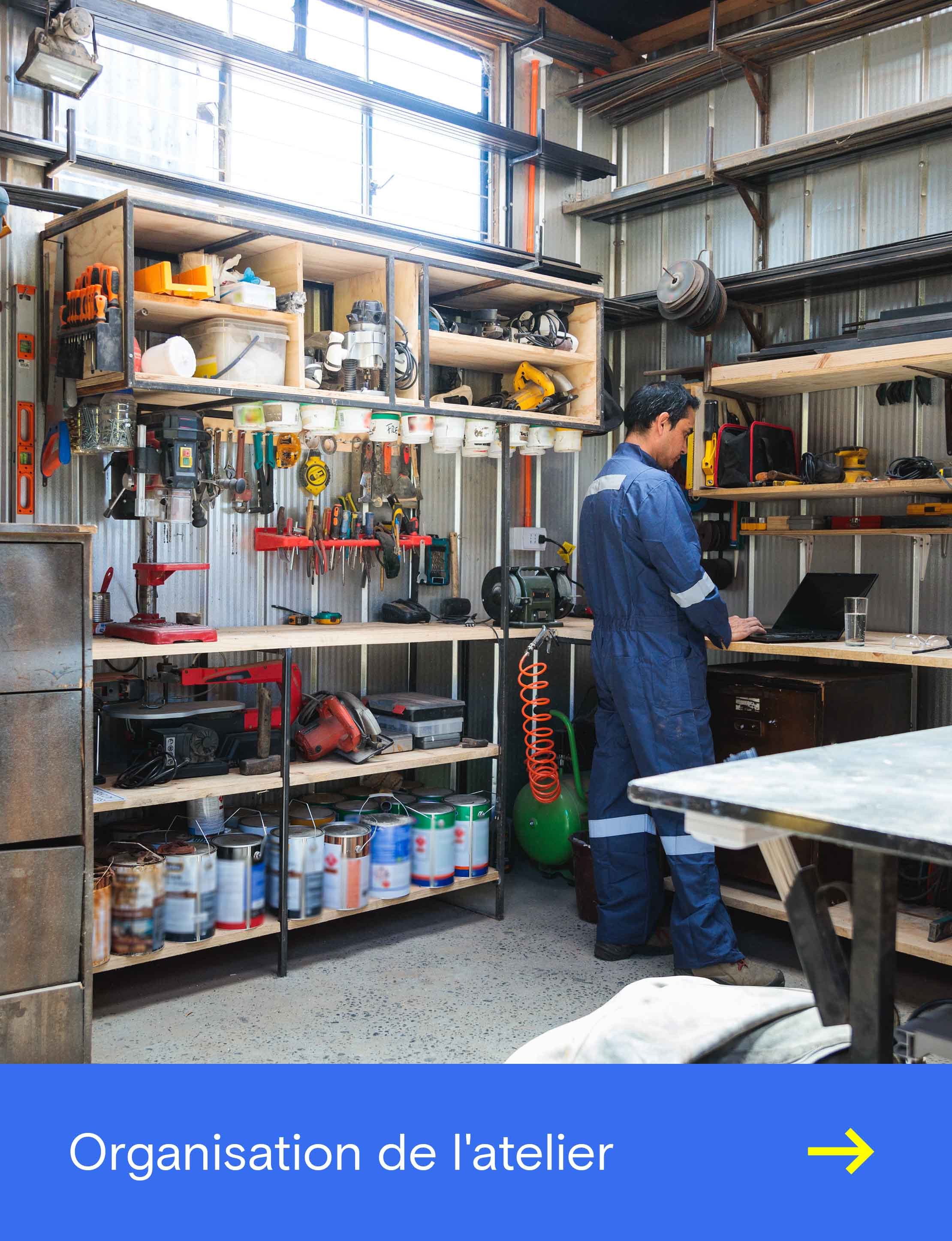 Un homme travaille sur un ordinateur portable dans un atelier bien équipé, entouré d’outils et de machines.