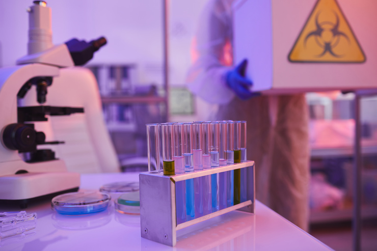 Le personnel de laboratoire vêtu de tenues de protection transporte un conteneur de substances dangereuses vers une zone sécurisée du laboratoire ; au premier plan, des appareils de laboratoire et des échantillons.