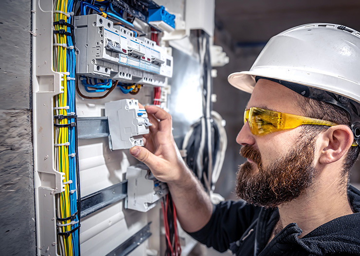 Un électricien, équipé d'un casque et de lunettes de protection, installe un disjoncteur dans un coffret électrique ouvert.