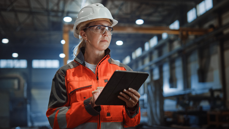 Une femme en tenue de protection et casque tient une tablette dans une usine.