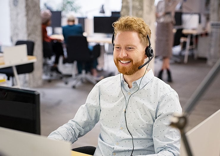 Une personne équipée d'un casque travaille en souriant devant un ordinateur dans un bureau moderne en open space.