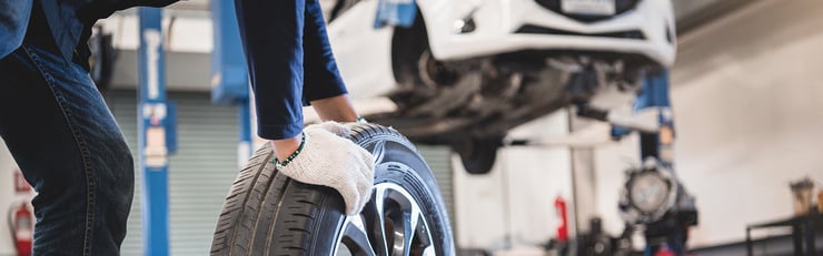 Une personne portant des gants de travail soulève un pneu de voiture dans un atelier, tandis qu'un véhicule est posé sur un pont élévateur.