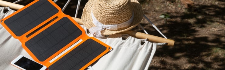 Panneau solaire pliable recharge un smartphone sur un hamac en extérieur, à côté d’un chapeau de paille.