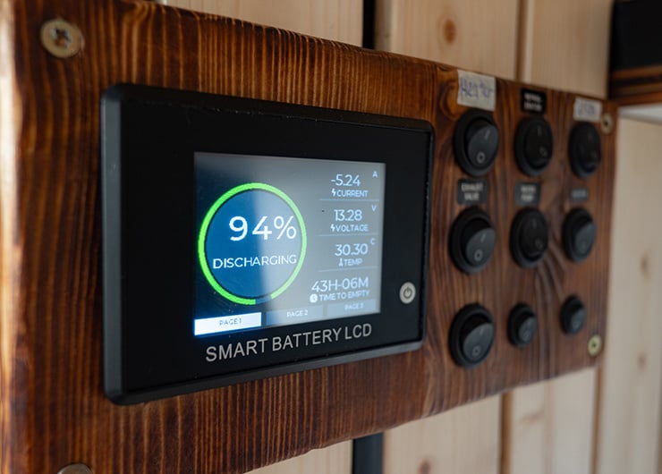 Un écran LCD intelligent affiche l'état de charge d'une alimentation électrique avec un affichage en pourcentage et des valeurs techniques mesurées sur un panneau en bois.