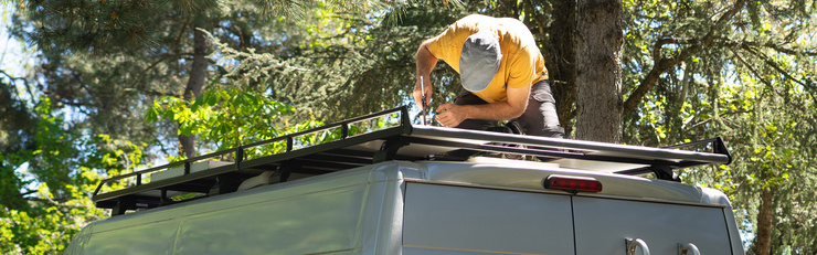 Une personne installe une galerie de toit à l'extérieur, sur le toit d'un véhicule, entourée d'arbres.