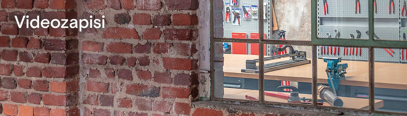 Pogled kroz stari metalni prozor u radionicu s alatima i rezačem pločica. Na zidu od opeke napisana je riječ 'Videos'.