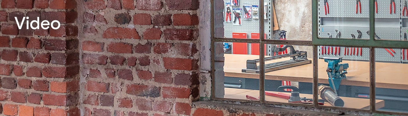 Een blik door een oud raam met metalen kozijn naar een werkplaats met gereedschap en een tegelsnijder. Op de bakstenen muur staat ‘Video's’.