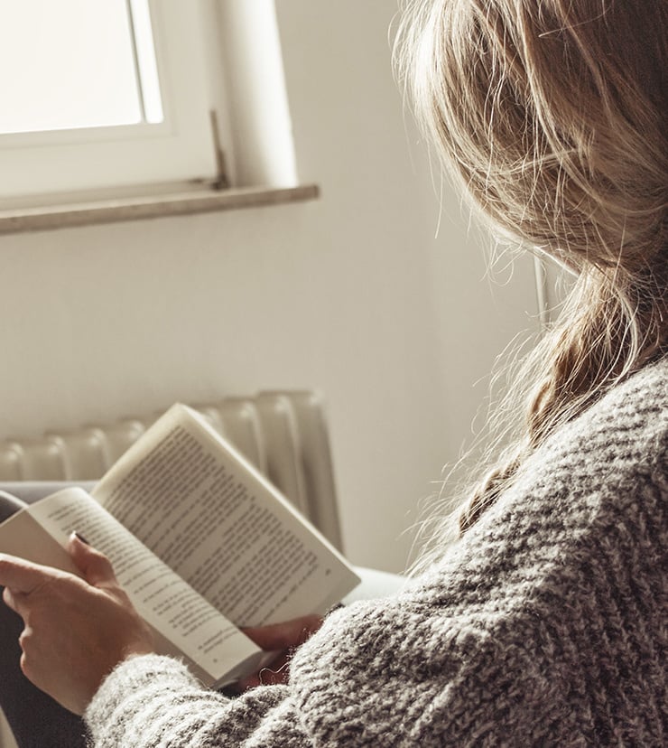 Een vrouw met licht haar leest een boek, met een trui met kabelpatroon, in een lichte kamer met een raam op de achtergrond.