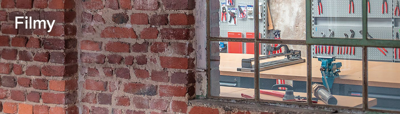 Widok przez stare okno z metalową ramą do warsztatu z narzędziami i przecinarką do płytek. Na ceglanej ścianie widnieje napis „Videos”.