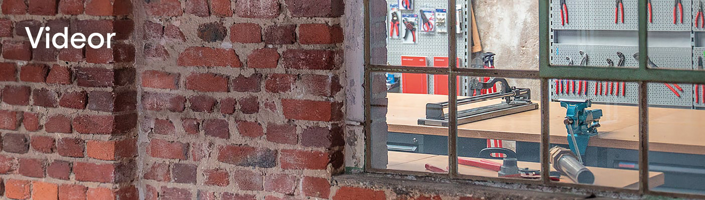 En vy genom ett gammalt metallinfattat fönster in i en verkstad med verktyg och en kakelskärare. Ordet "Videor" står skrivet på tegelväggen.